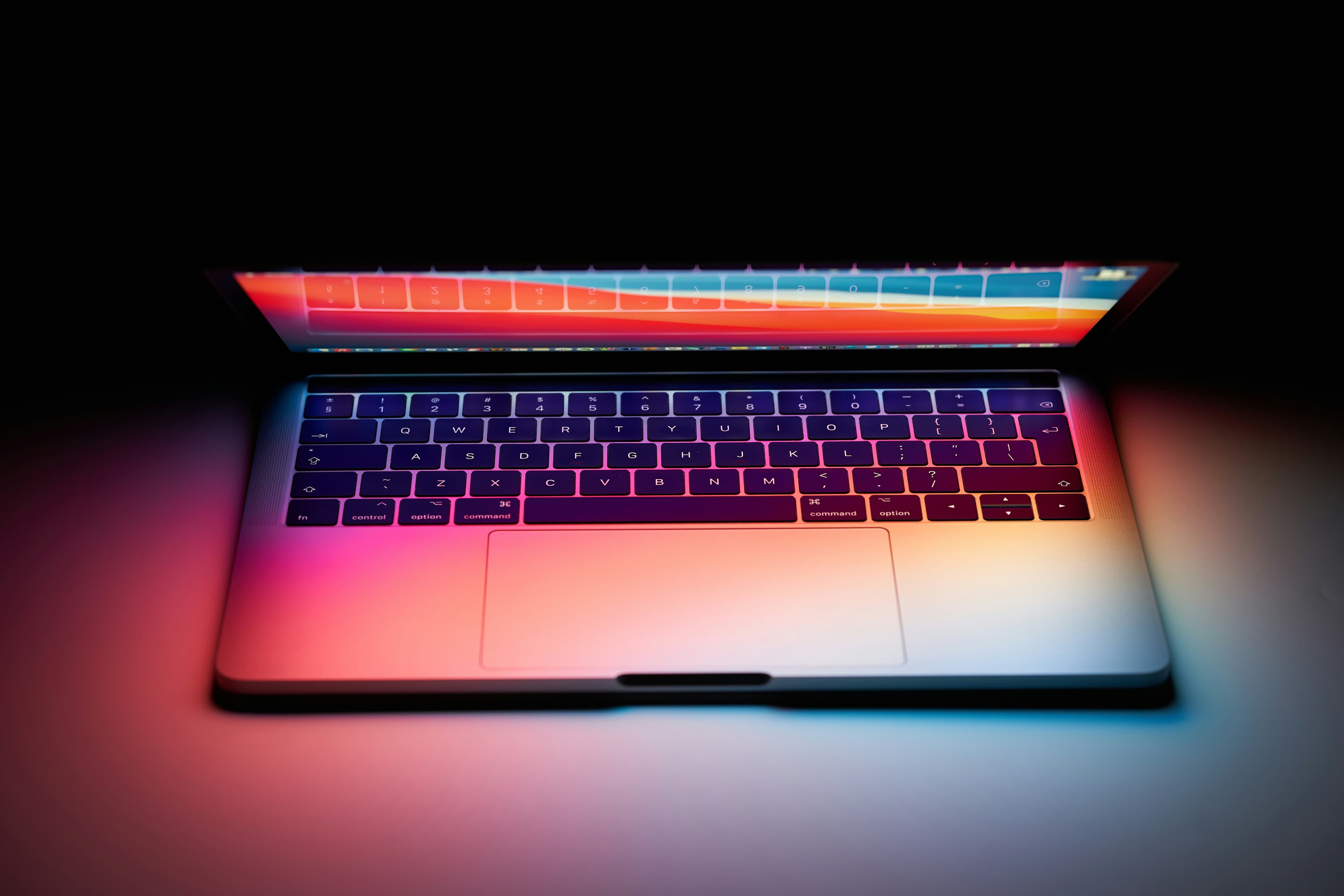 A MacBook with a colourful wallpaper glowing in a dark room